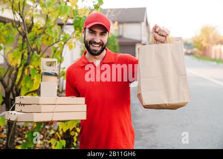 Image of smiling young delivery man in red uniform holding pizza boxes and paper bag at city street Stock Photo