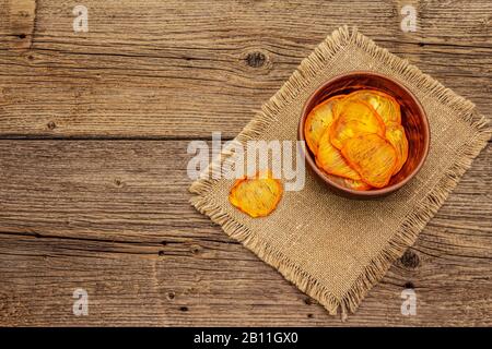 Dried slices of persimmon in bowl. Fruit snack, healthy eating concept. Sackcloth, old wooden boards background, top view, copy space Stock Photo