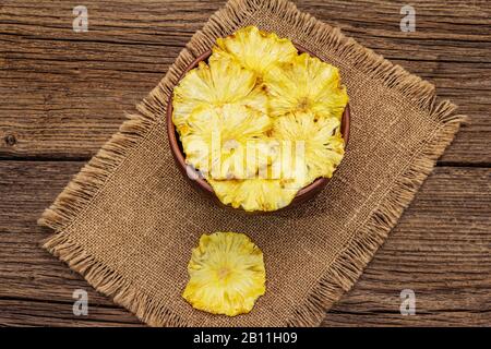 Dried slices of pineapple in bowl. Fruit snack, healthy eating concept. Sackcloth, old wooden boards background, top view, copy space Stock Photo