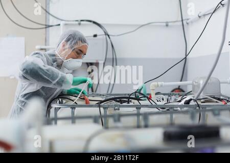 Young man in protective clothing working with wires during epidemic in the plant Stock Photo