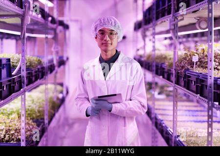 Portrait of Asian young botanist in white coat using tablet pc and looking at camera while working in greenhouse Stock Photo