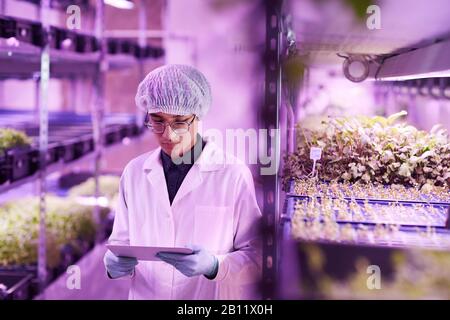 Asian young man in white lab coat working online on digital tablet while standing in greenhouse Stock Photo