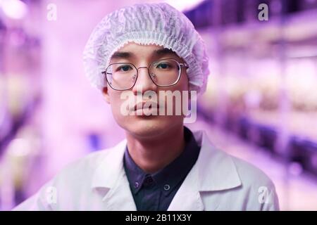 Portrait of Asian young scientist in eyeglasses and in cap looking at camera while working in the lab Stock Photo