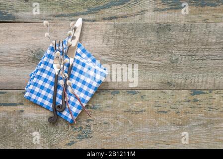 Easter serving in vintage style: forged handmade knife and fork, two fluffy willow twigs and checkered blue-and-white napkin lie on a rough wooden bac Stock Photo