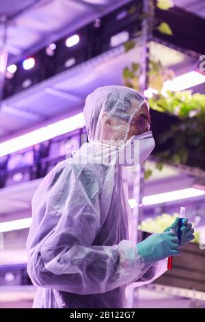 Young botanist in protective clothing holding test tubes while working in greenhouse Stock Photo