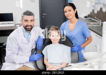 Happy little girl in armchair with dentist and his assistant standing near by Stock Photo