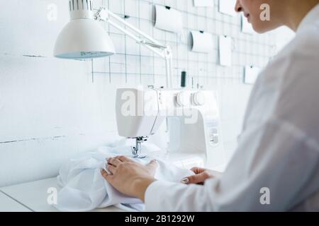 Young female sewing factory worker in white workwear sitting by electric machine Stock Photo