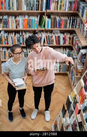Two casual teenagers choosing books from bookshelf in college library Stock Photo