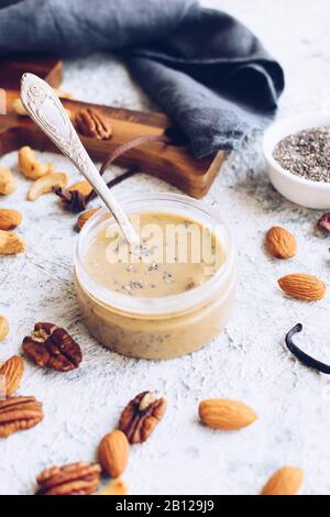Almond, pecan and cashew nut butter in glass jar. Homemade raw organic mixed nuts paste with chia seeds and vanilla on grey background. Healthy natura Stock Photo