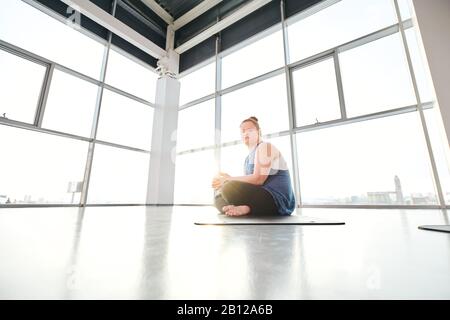 Young disable female with crossed legs doing yoga exercise on mat in gym Stock Photo