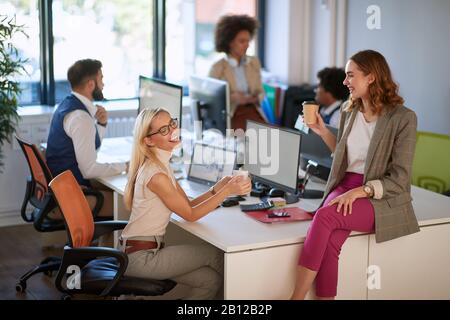 two female colleagues talking, drinking coffee and smiling at work, with group of colleagues in the background. casual, open space, modern, office, bu Stock Photo