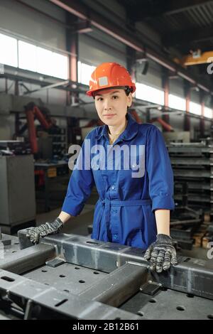 Pretty young confident factory worker in blue uniform, gloves and helmet Stock Photo