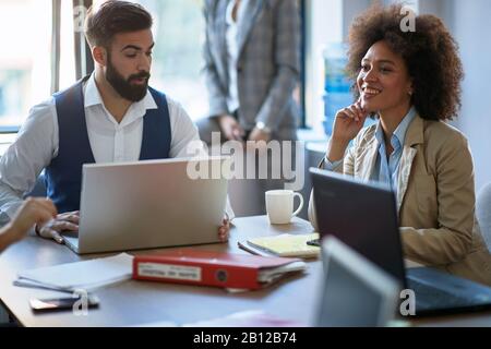 two coworkers sitting at the desk in office with laptops, talking, exchanging information Stock Photo