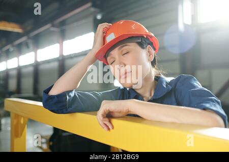 Tired young female engineer in workwear and hardhat napping by bar Stock Photo