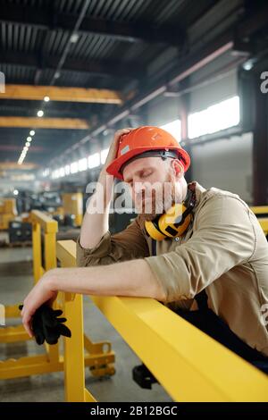 Tired or sick engineer in workwear and hardhat leaning by bar while having break Stock Photo