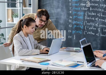 Clever student sitting by desk in front of computer while teacher checking data Stock Photo
