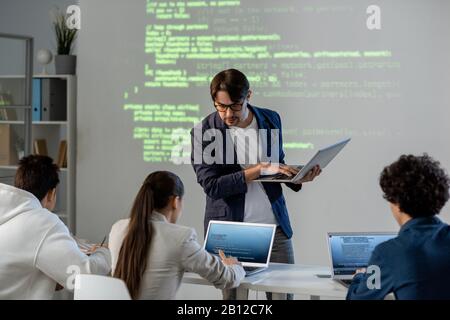 Teacher with laptop standing by desk and bending over one of students Stock Photo