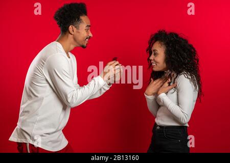Young couple. African man makes marriage proposal to his lover woman with ring on red studio background. Love, holidays, happiness concept. Stock Photo