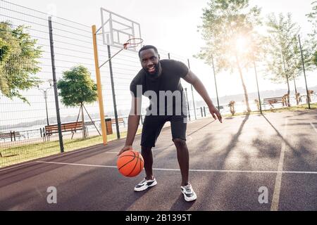 Outdoors Activity. African man playing basketball on court dribbling smiling excited Stock Photo