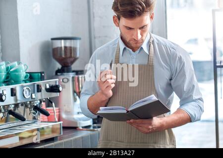 Coffee Passion. Young barista at coffee shop reading instructions at book concentrated Stock Photo