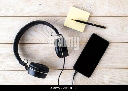 Smartphone, black headphones and pen with sticky notes notes block on a white wooden table. Personal device with touchscreen for music and communicati Stock Photo