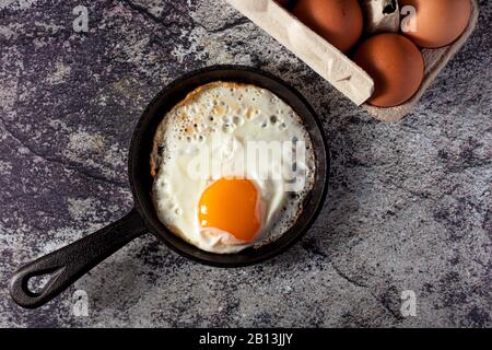 Sunny side up fried egg in cast iron skillet with whole eggs on concrete background. Top view Stock Photo