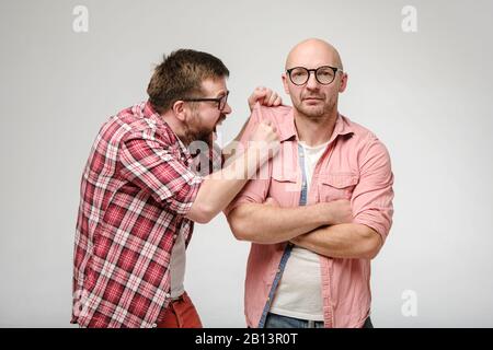 Angry bearded man screams furiously and grabs his friend shirt sleeve, who stands unperturbed with arms crossed. Stock Photo