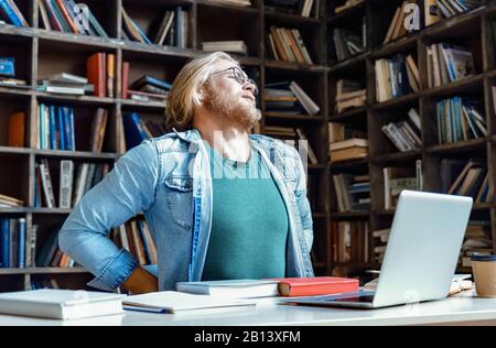 Tired upset young man glasses stretch library desk feel back pain concept Stock Photo