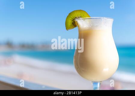 An ice cold mixed liquor drink with a fresh kiwi slice on the rim in front of a tropical beach and sea. Selective focus shot on the kiwi. Stock Photo