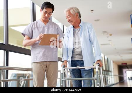young asian physical therapist working with old man on working using a walker in hallway of nursing home Stock Photo