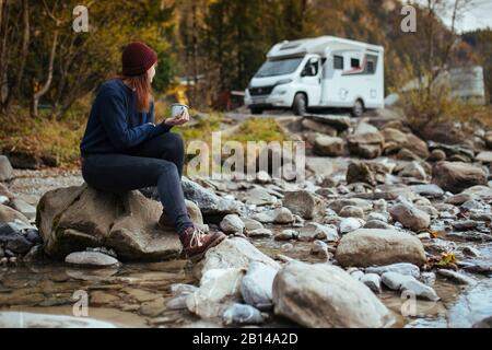 Road trip l woman sits with cup of tea in front of motorhome Stock Photo