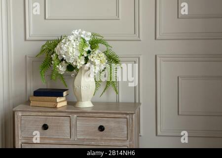 A white vase with white flowers stands on a dresser against the wall. Nearby are three books. Classic interior. Stock Photo