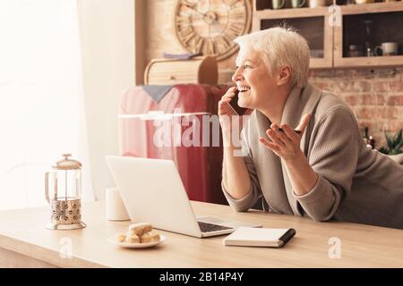 Cheerful elderly woman talking on cellphone and using laptop in kitchen Stock Photo