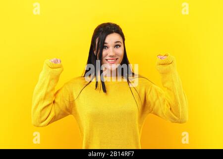 brunette girl rejoicing her success and victory clenching her fists with joy. Lucky woman in a yellow sweater smiling at the camera Stock Photo