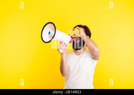 Happy bearded hipster man with sliced lemon on eyes, shouting through a megaphone, over yellow background, concept of summer mood Stock Photo