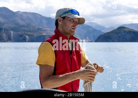 A man on a yacht is tying a knot on a rope, wearing a red yolk, wearing a cap and glasses. Stock Photo