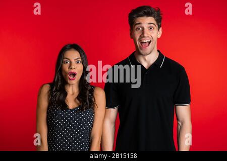 Amazed european man and woman shocked, saying WOW. Young couple in black surprised to camera over red background. Stock Photo