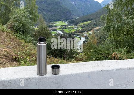 Thermos on concrete, drinking tea on green and rocky Norway hills. Water running down, small village in northern mountains landscape. Nature travel sc Stock Photo