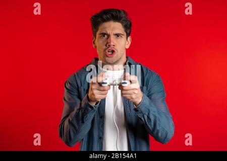 Handsome young man playing video exciting game on Tv with joystick on red studio wall. Using modern technology. Stock Photo