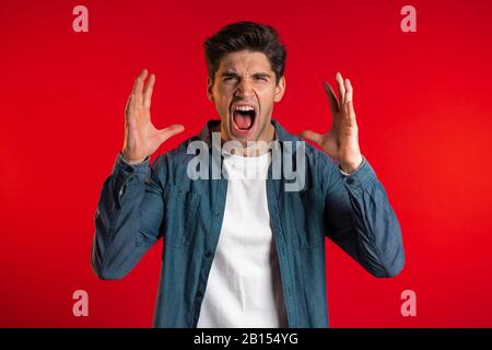 Young european stressed man in blue shirt shouting isolated over red background. Depressed guy loudly screaming to camera. Stock Photo