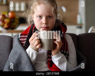 portrait of a sick child in a scarf and plaid with a cup of hot tea on the sofa in the apartment Stock Photo