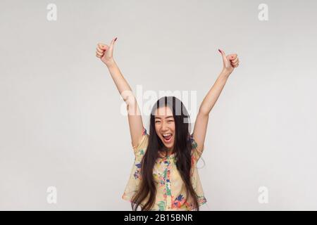 I did excellent job! Delighted pretty asian girl in summer blouse shouting in excitement, raising hands and showing thumbs up, emotionally reacting to Stock Photo