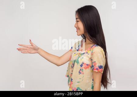 Side view of friendly asian girl with long brunette hair in blouse giving hand to handshake and smiling to client, greeting and getting acquainted. in Stock Photo