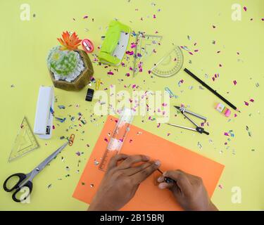 Craft materials scattered on a light green paper background with sparkle paper,while a person is making a circle on a orange paper Stock Photo