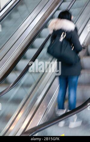 Young teenager woman in movement standing alone in an urban up moving escalator leaving  the subway station traveling upstairs to the city mall. Stock Photo