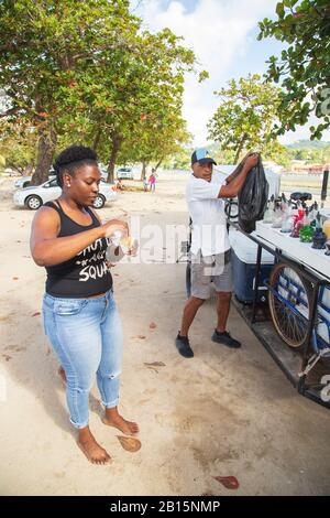 Young black lady with natural hair eating ice-cream on the beach on a hot day while ice-cream vendor watches her while packing a black nylon bag Stock Photo