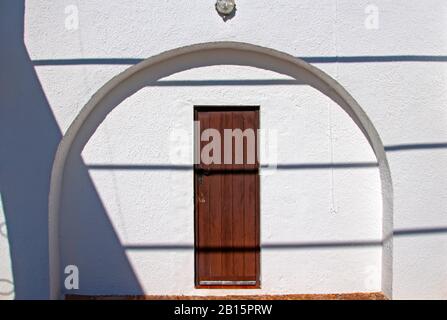 Brown wooden door in the arc on the white wall with shadows. Summer, Spain Stock Photo
