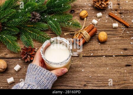 Christmas eggnog liqueur or cola de mono cocktail. Classical winter drink in glass mug, xmas decorations. Woman's hand in jersey. Old wooden backgroun Stock Photo