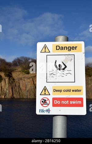 Warning sign Prestonhill quarry Inverkeithing where deaths have occurred. Deep cold water, danger of drowning, do not swim. Blurred water, cliffs. Stock Photo