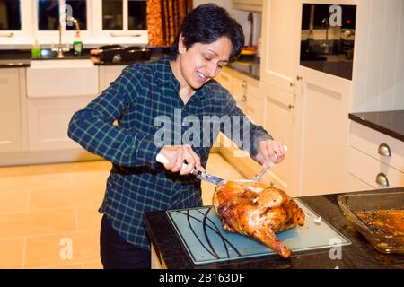 Asian woman carving meat with a knife and fork in a home kitchen for a roast turkey meal, UK Stock Photo